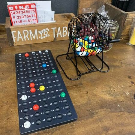 a black wood bingo board and a bingo ball hopper on a dooen table with a wood box that says Farm to Table in the background that is holding bingo cards