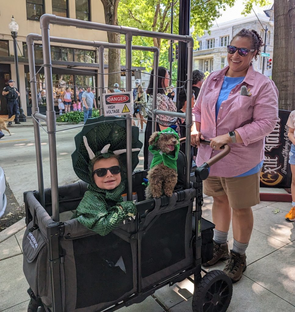 woman with a wagon made to look like a dino cage with a kid and a dog dressed as dinosaurs inside