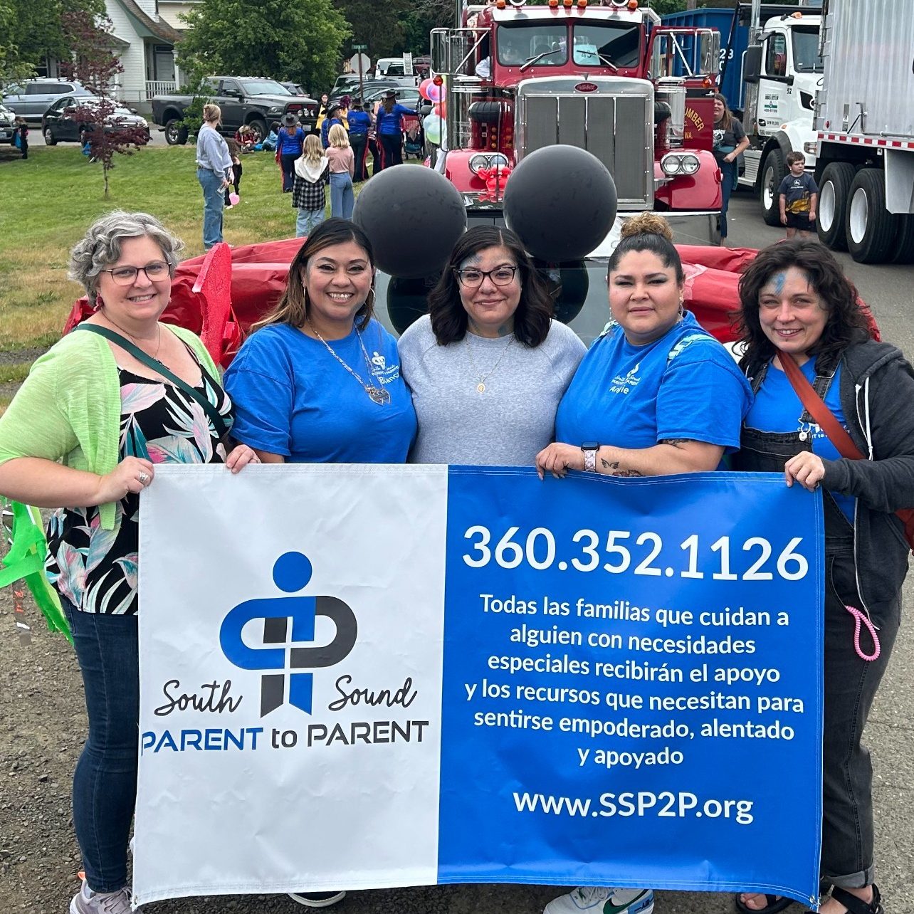 five woman post in front of a fire truck with lots of people milling around. They are holding a banner that says 'south sound parent to parent. 360.3562.1126 with text in spanish and a website: www.sspsp.org
