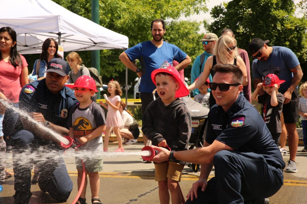 a small child helps hold a fire hose with a fireman while it shoots water
