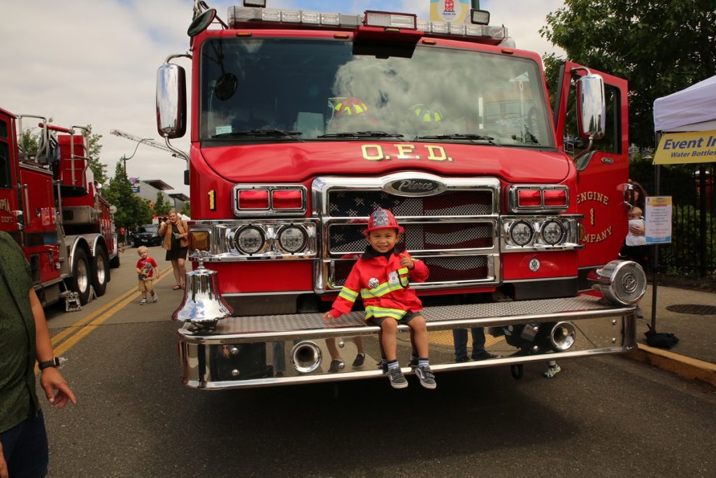 little kid in a firefighter's coat sitting on the front bumper of a fire truck.,