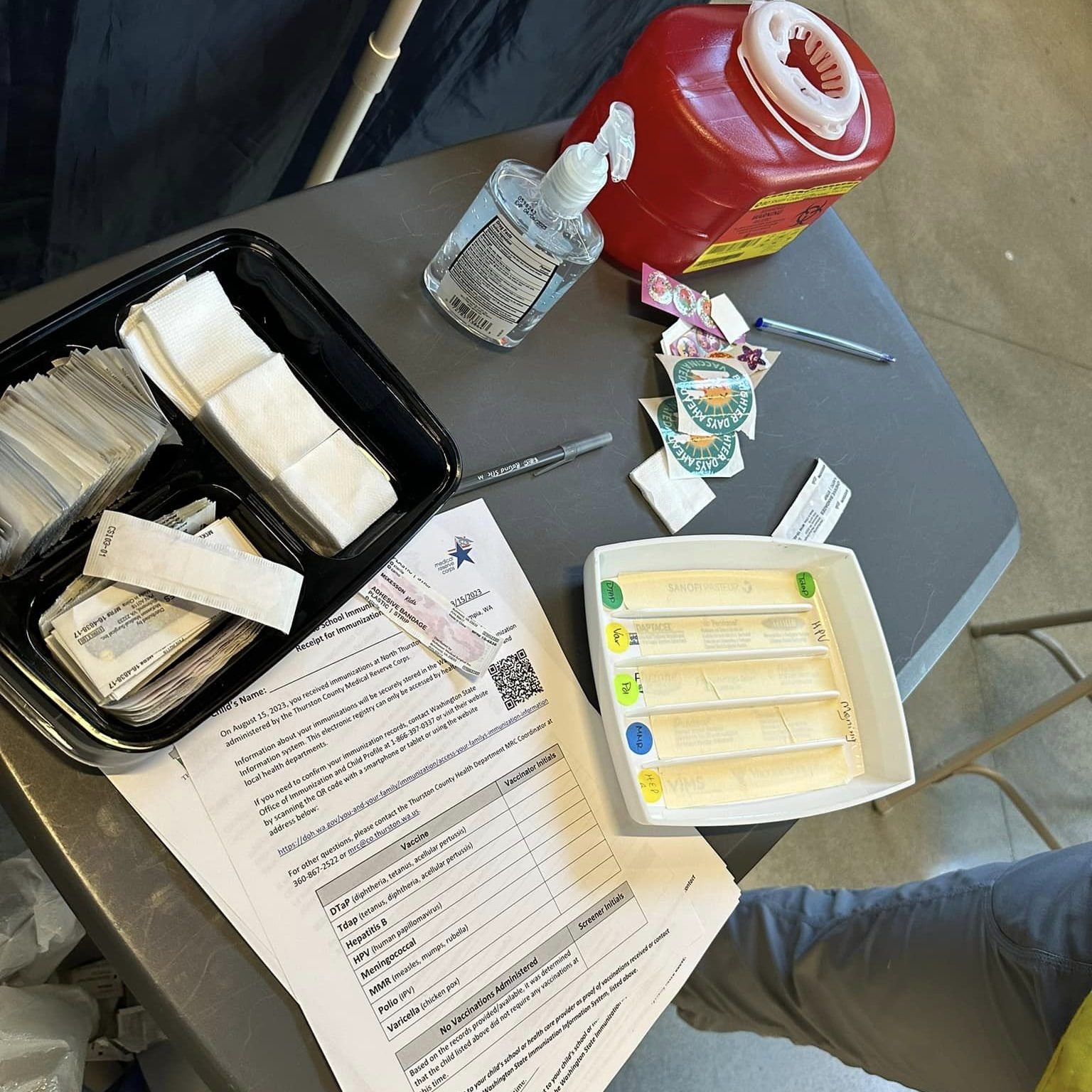 table with vaccine items on it including sharps container, sanitizer, cotton pads, band aids and paper forms