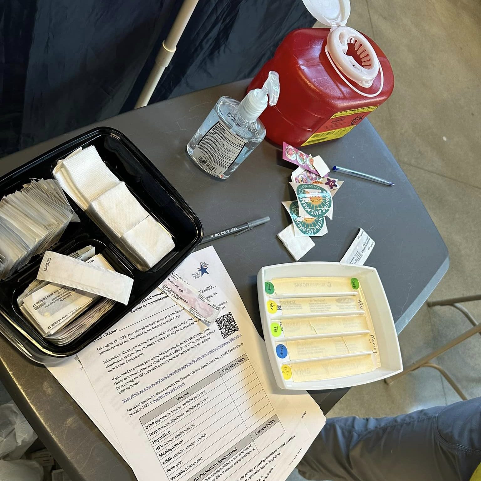 table with vaccine items on it including sharps container, sanitizer, cotton pads, band aids and paper forms