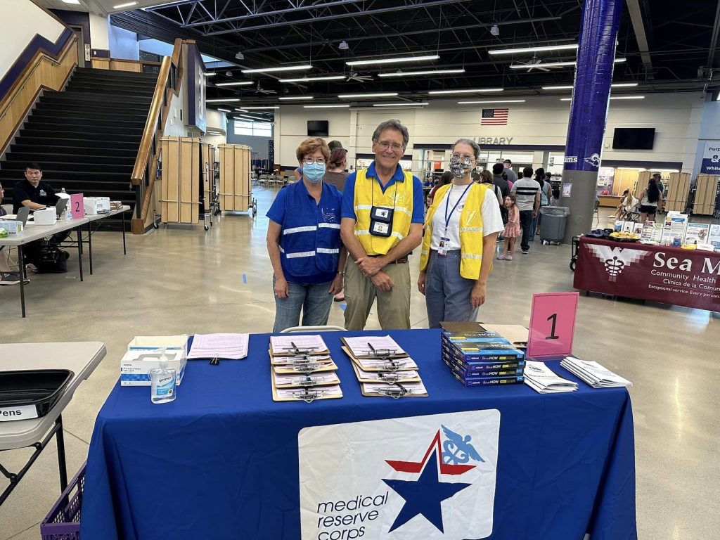 three people standing behind a table in an expo center. Table has a blue cloth and pamphlets on it.