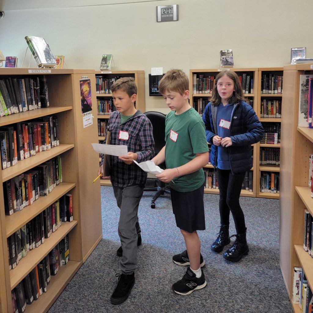 three boys with name tags, two of them holding pieces of paper, in the NOVA library