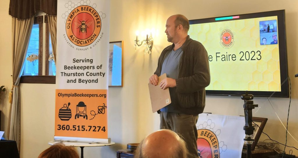 a man giving a talk in a room with a banner that says Olympia Beekeepers Association