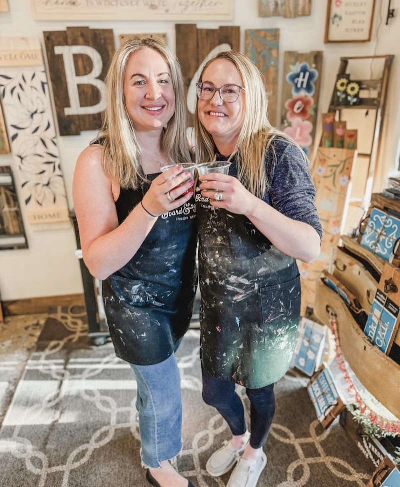 Two woman wearing aprons covered in paint, pose for a photo holding to classes