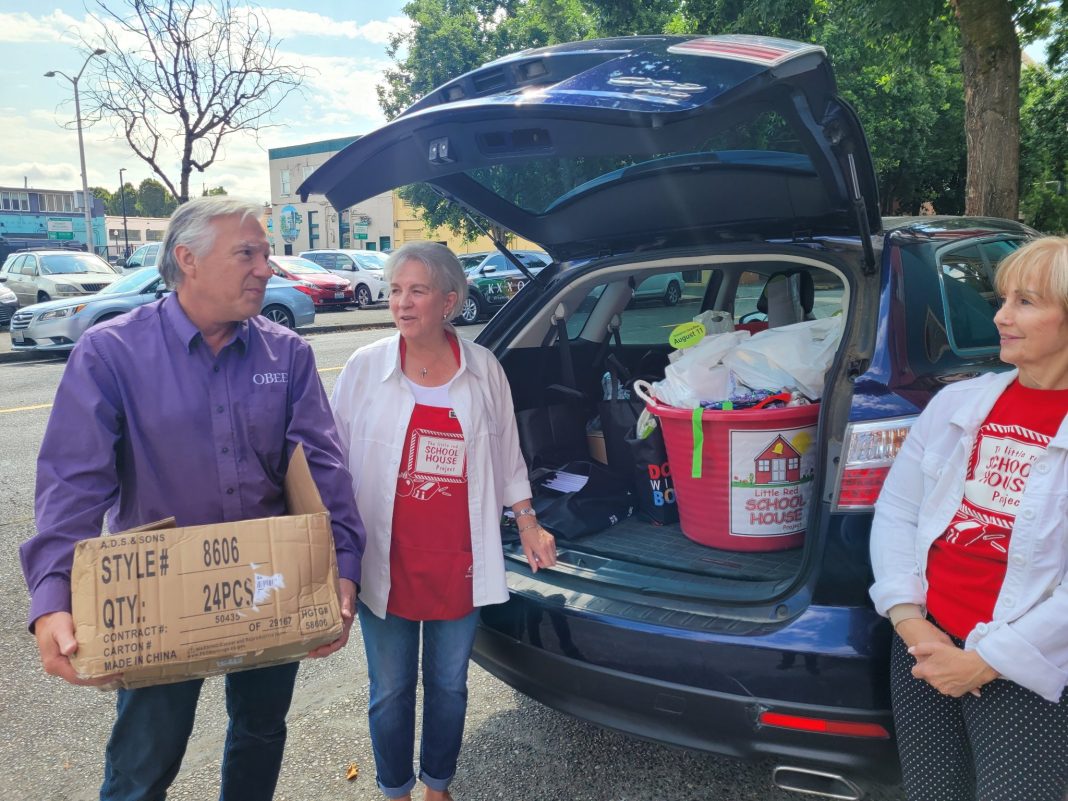 People standing next to car trunk that is holding school supplies. One man is carry a large cardboard box