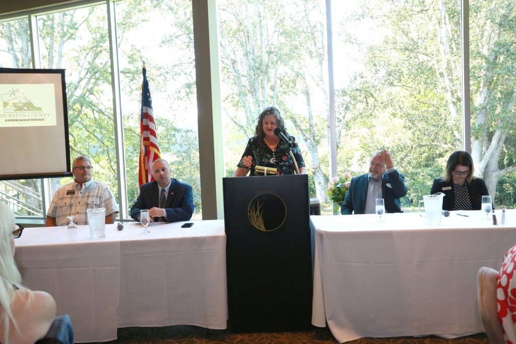 a woman stands a podium, speaking, with long white tables with people seated at them on either side of her