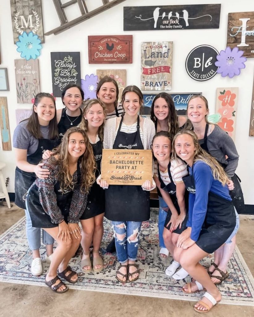 a group of women pose in front of a wall of wooden signs. One in holding a wood design that says 'I celebrated my bachelorette party at Board & Brush'