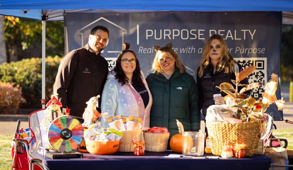 Two people with their faces painted and two without stand behind a table full of fall decorations and treats under a pop up tent