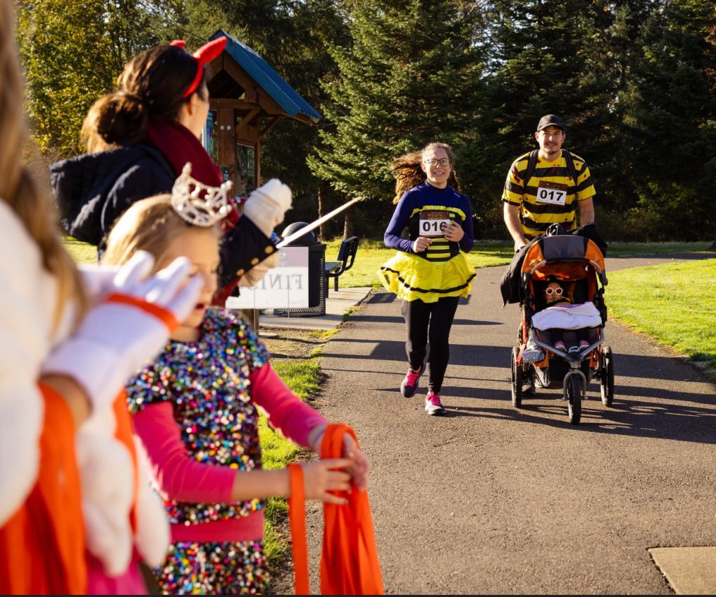 people in bee costumes, one pushing a stroller, run down a concrete path at a park with people in costumes in the foreground