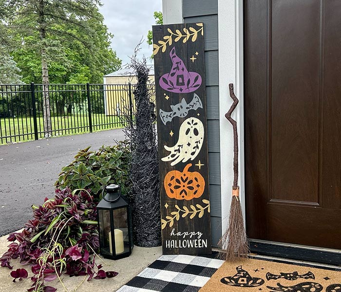 wooden board painted with a witch hat, bat, ghost and pumpkin by a door