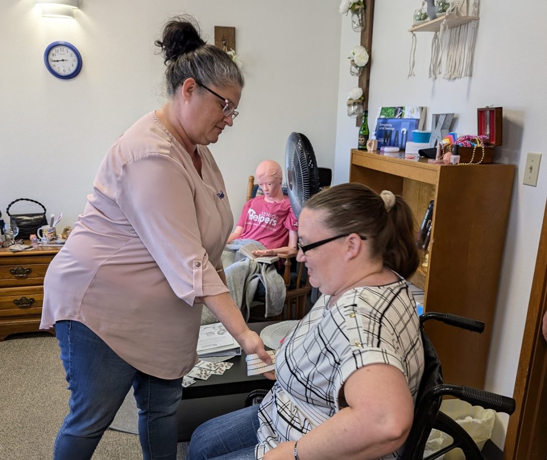 a woman helps a woman in a wheelchair with something, a mannequin is sitting on a chair next to her