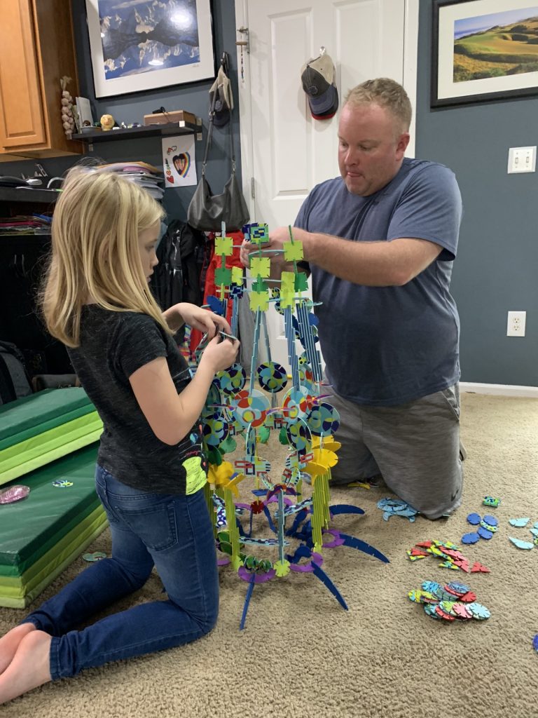 a child and her father knee on carpet building a tall tower out of some type of plastic pieces