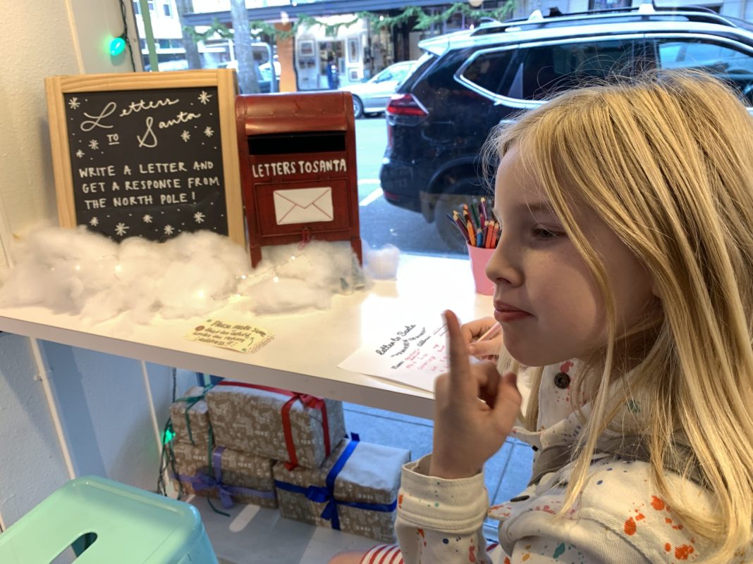 a girl puts her finger up to her mouth while writing a letter to Santa at a counter with a little red mail box on it