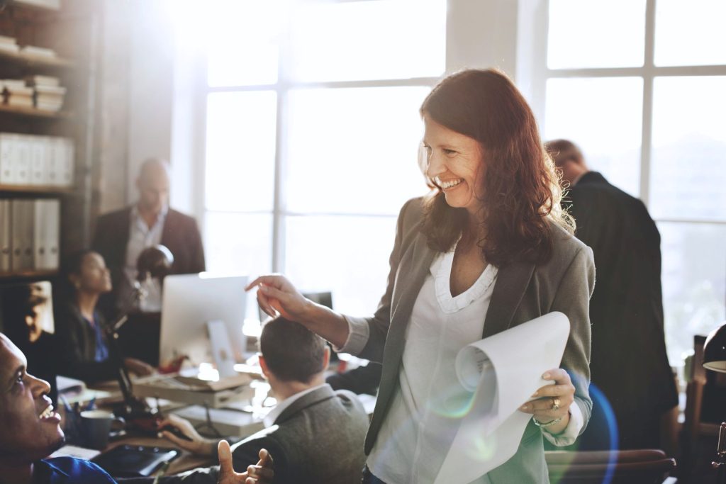 a woman standing up smiles at a guy sitting. In the background at other people sitting at a desk with desktops and laptops