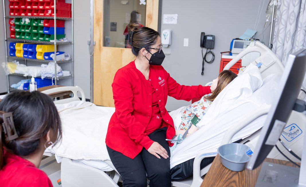 Saint Martin's University Nurse student in red scrub top and face mask sits on the edge of a hospital patient with her hand by fake mannequin patient's head