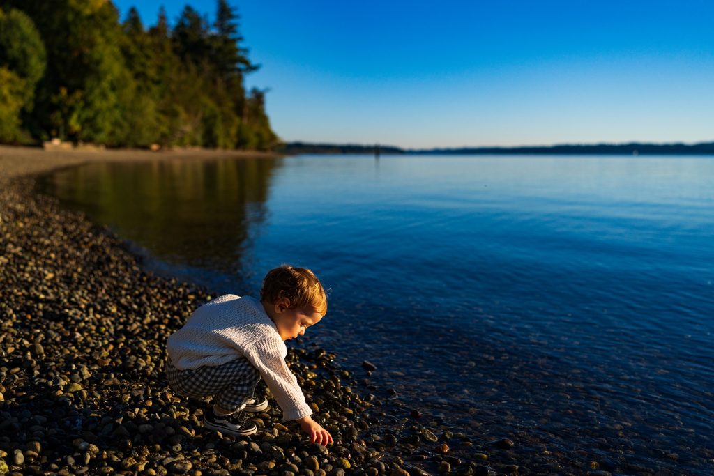 a little boy crouches to pick up a rock on a shoreline