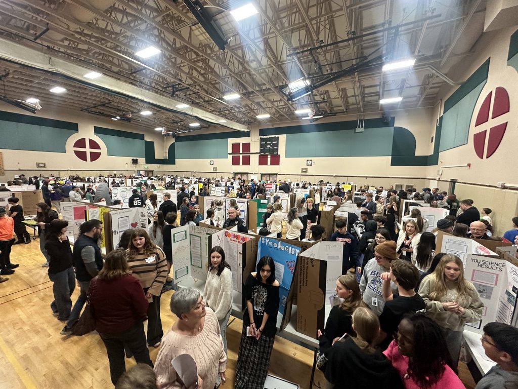 large group of people in a middle school gym walk around and look at projects boards