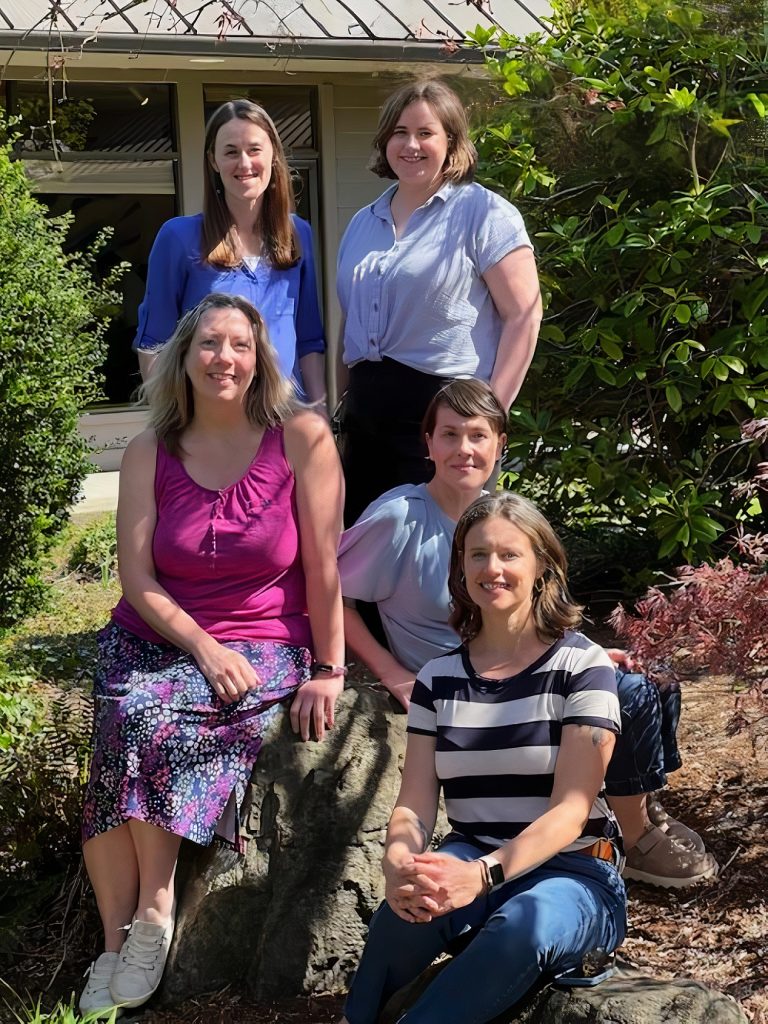 five women smile for the camera, two seated on a rock, one crouched in front of it, the other two standing behind it
