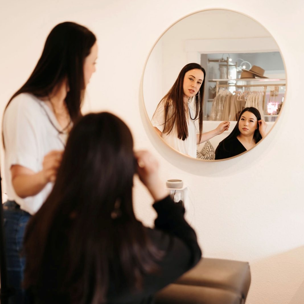 a person in a salon chair talks to a stylist standing next to them, both are looking in a round mirror