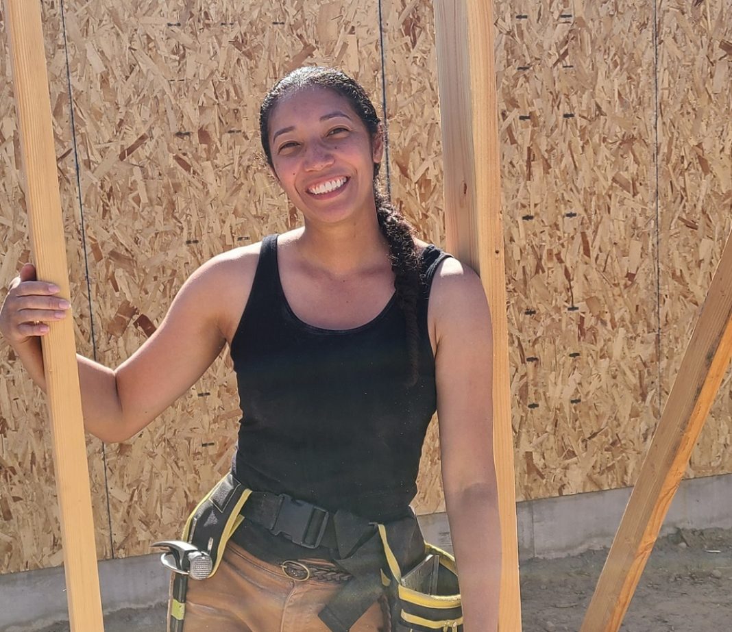 Kim Parks posing for a photo between framing on a house being built, wearing a black tank top and a tool belt