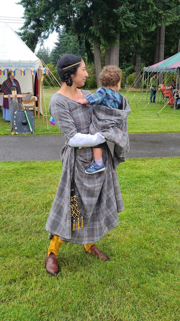 Kim Parks holding a child. Both are in matching medieval outfits. There are large open tents with people behind her in the grass