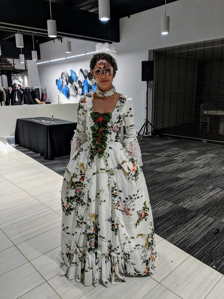 Kim Parks wearing a renaissance style ballgown and a masquerade mask inside a building with a black clothed table behind her.