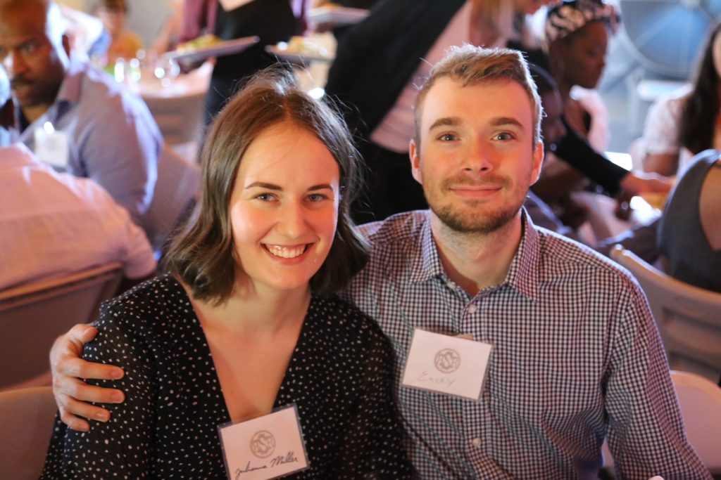 Juliana (left) and Emery (right) sitting together at a table, Emery has his arm around Juliana's shoulders