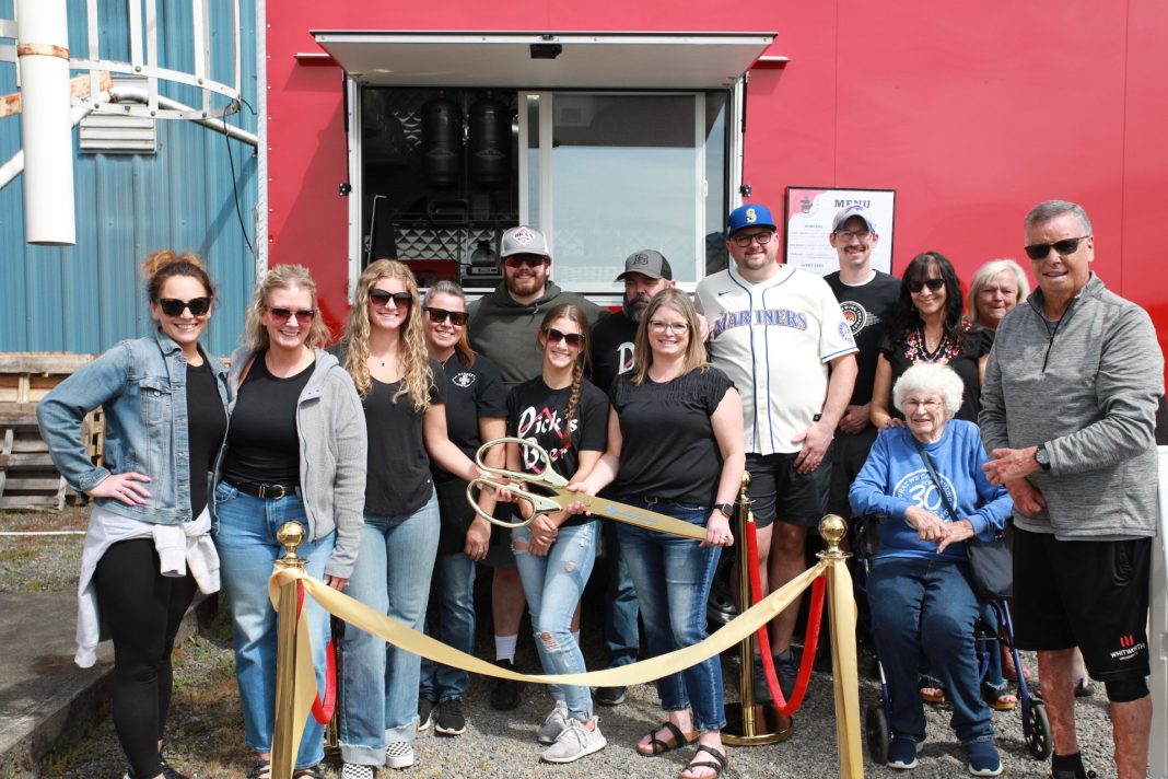 a large group of people who work at Dick's Brewing Company standing by a gold ribbon. A woman in the middle holds a giant pair of gold scissors