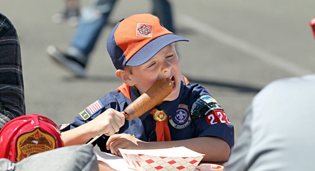 kid in a cub scout uniform eating a corn dog at a table