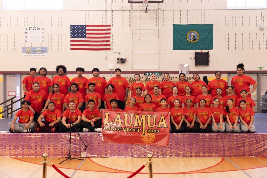 Laumua Youth Samoan Dance Group sitting on bleachers in red team shirts and black shorts with a banner that says Lauma Dance Group
