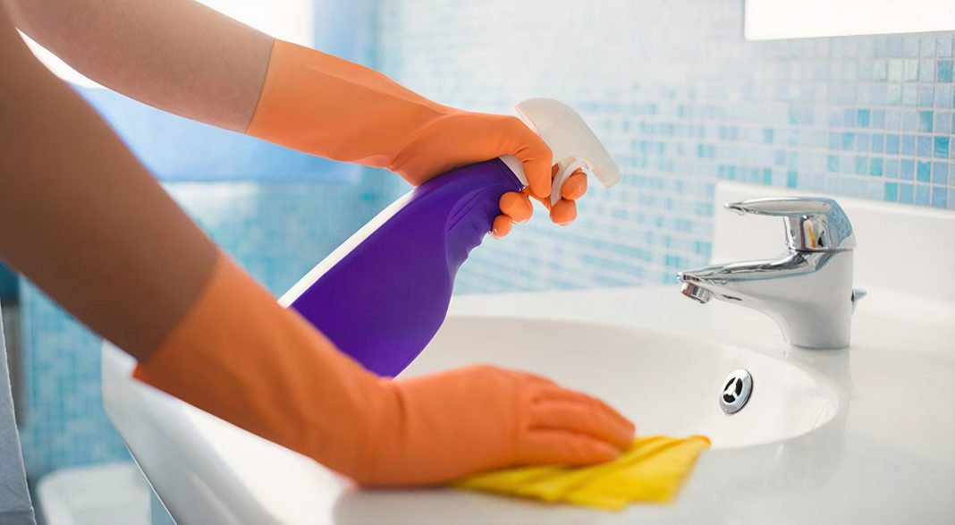 two hands with orange gloves, one has a yellow cloth on a white sink and the other is spraying something out of a purple spray bottle into the sink