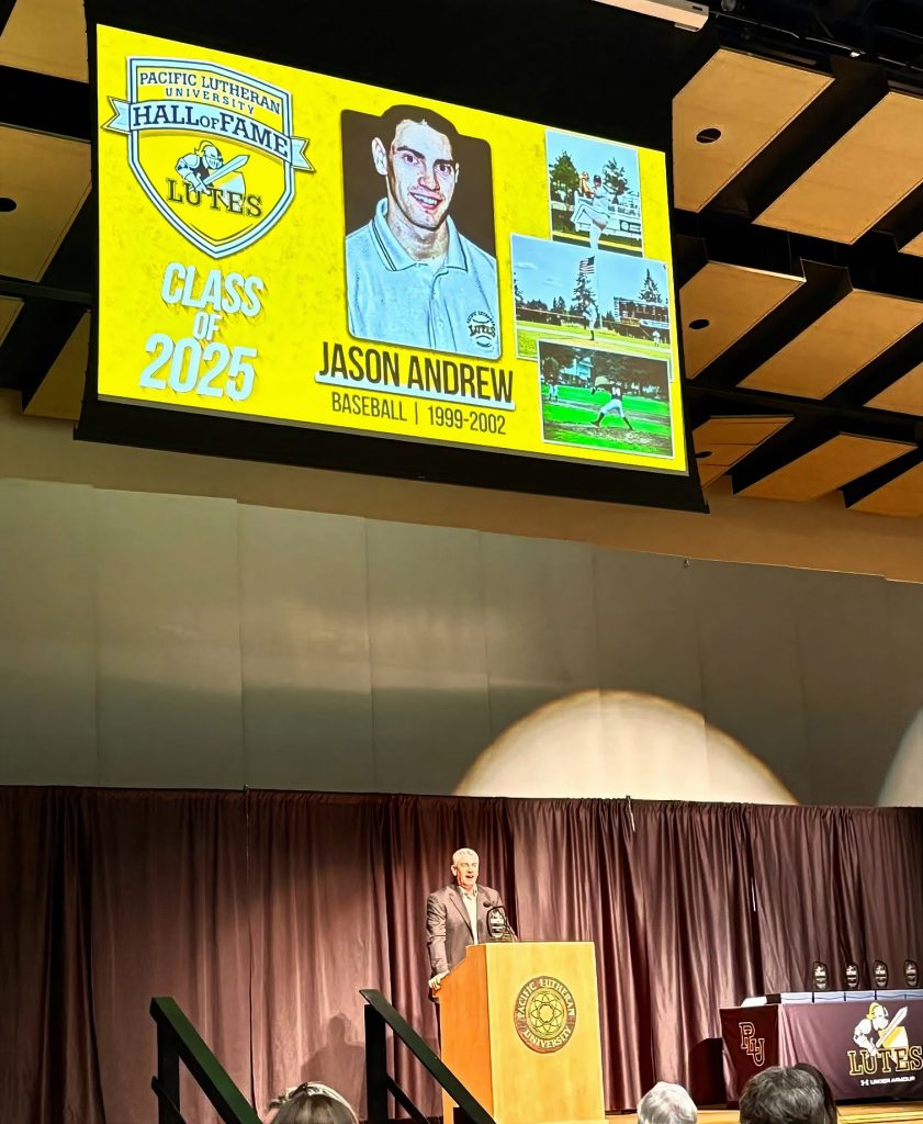 Jason Andrew in a suit standing at a podium with a Pacific Lutheran University logo. Above his head his a big screen with photos of him playing basseball as well as a headshot, with the words Jason Andrew, baseball, 1999-20002. Class of 2025. A logo in the top left says Pacific Lutheran University, Hall of Fame, Lutes
