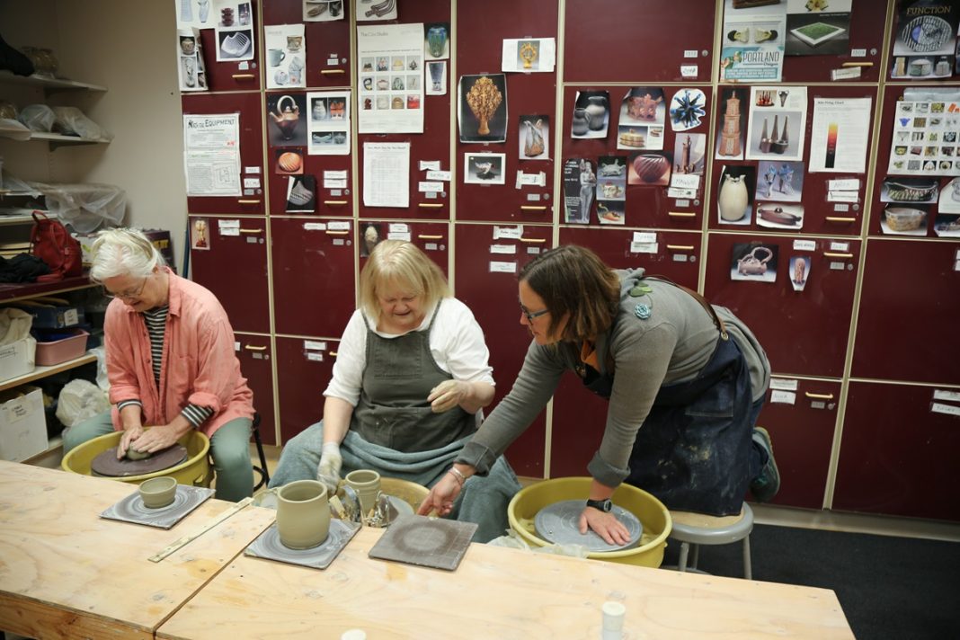 Two women working on potter's wheels at a table in a class through Olympia Parks, Arts, and Recreation. A third woman leans over and points at one of the woman's wheels