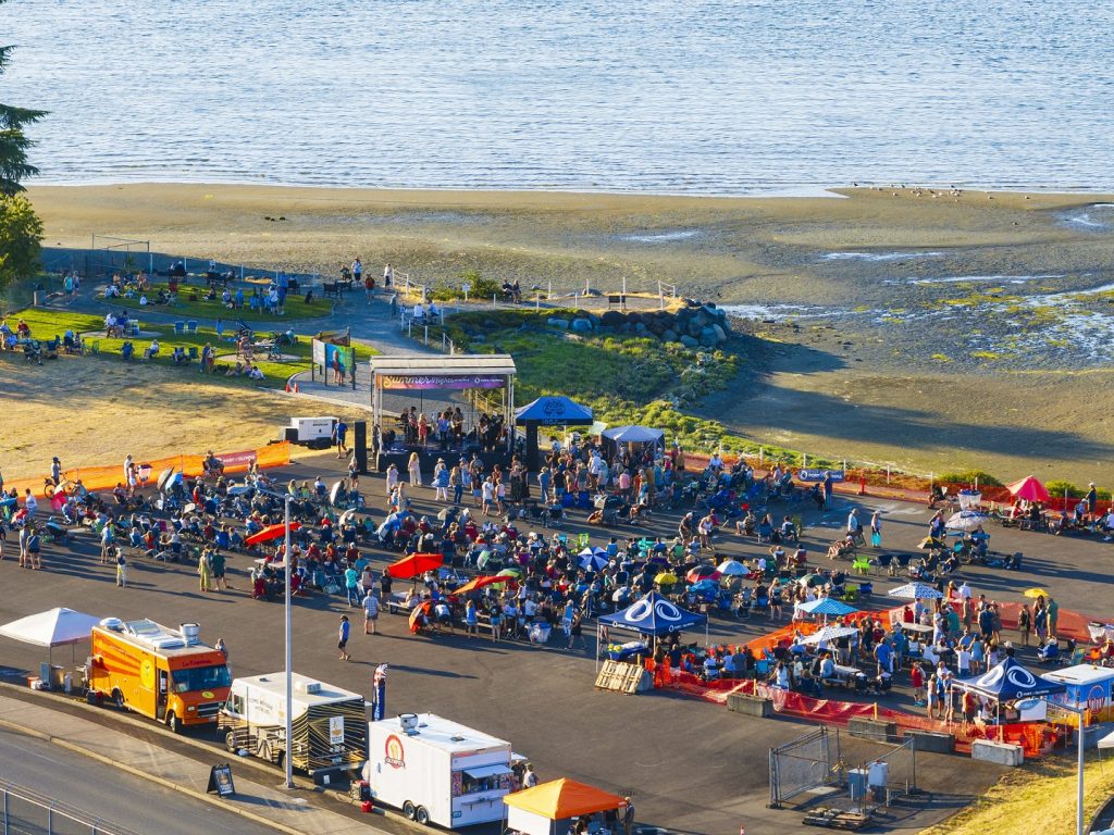 aerial photo of the Port of Olympia with huge crowds still in a parking lot with umbrellas, food trucks, pop up tents and a band on stage. The beach and water can be seen in the background