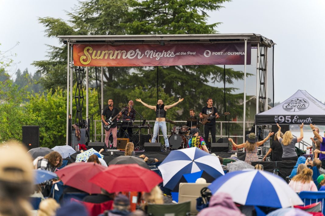 a huge crowd, some with umbrellas, watch a band perform on a large stage at Port of Olympia