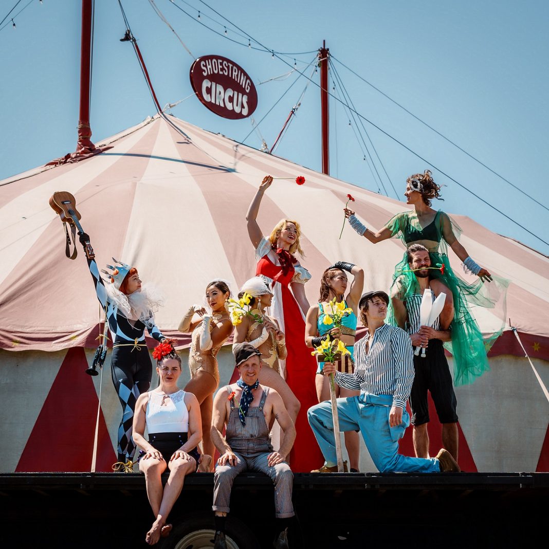 Circus performers in brightly colored costumes pose on a black stage in front of a red and white big top tent. Two are sitting, one is down on one knee, five are standing and one is standing with a woman on his shoulders.