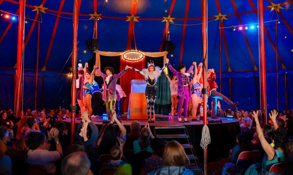 many ciricus performers in different costumes stand on a round stage with their hands linked and arms raised in front of a crowd