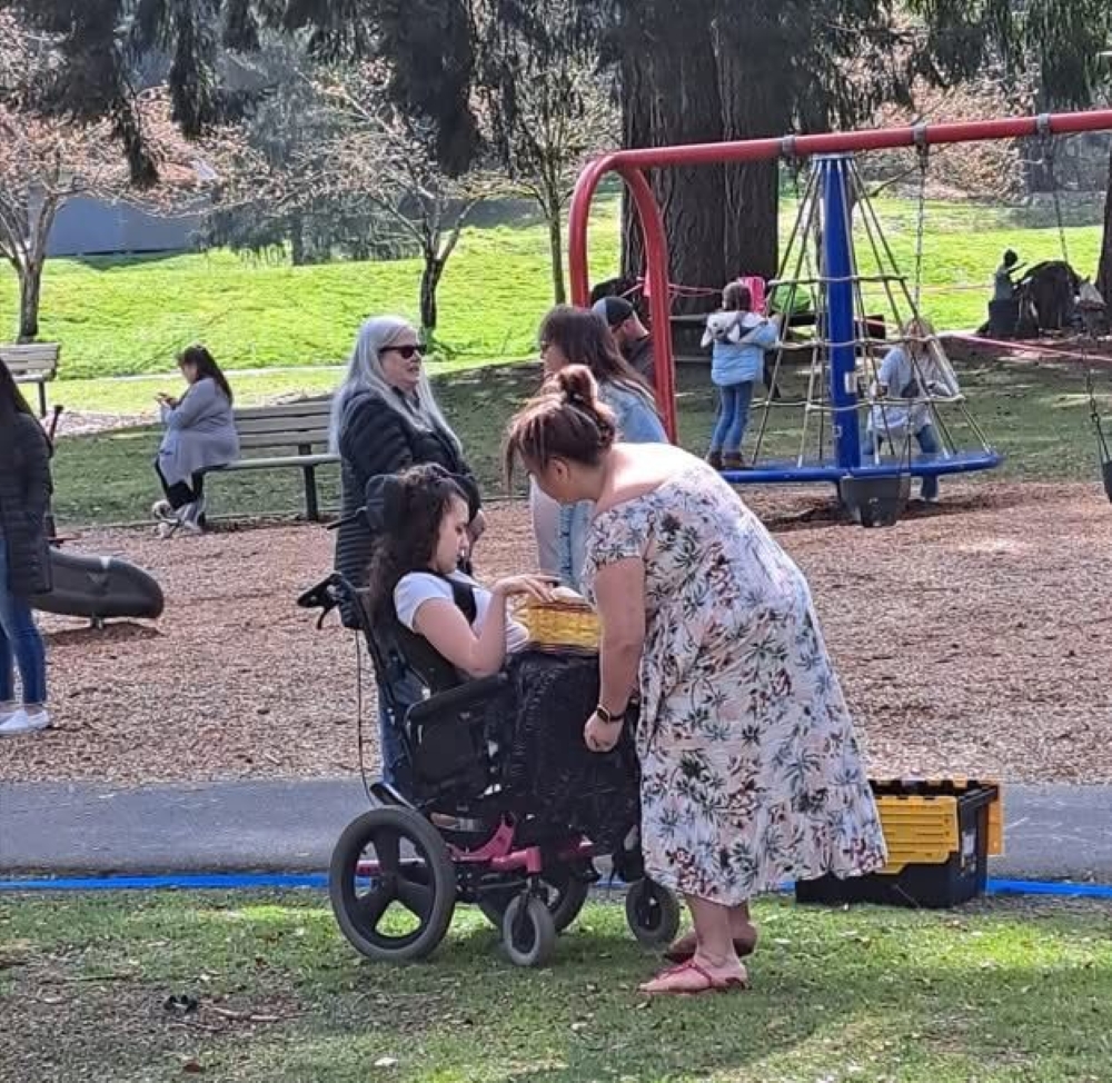 a woman leans over an older youth in a wheelchair, there are other adults and kids in a playground in the background
