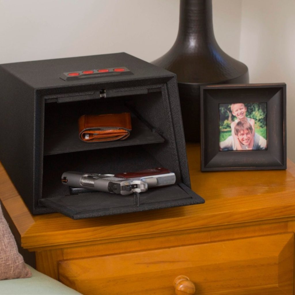 A small gun safe open with a gun insdie on a bedside table. There is a lamp and a photo of a guy with a kid on his shoulders next to it