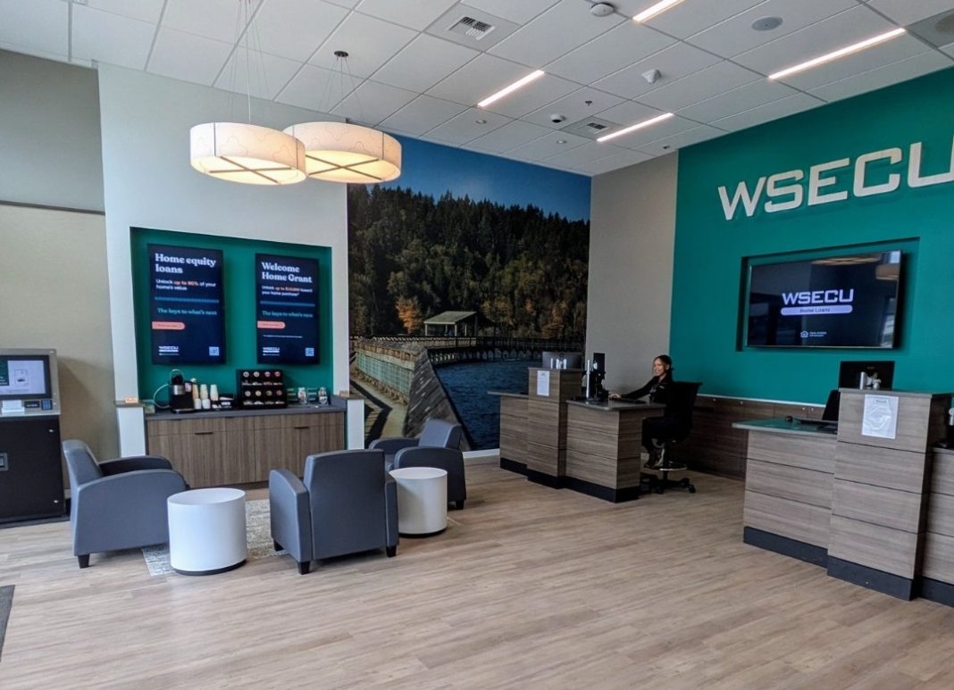 grey sofa chairs, white round side tables and tan cabinets in the WSECU Hawk's Prairie branch waiting area. The wall has a green block painted on it that has the word 'WSECU' painted on it in lighter green