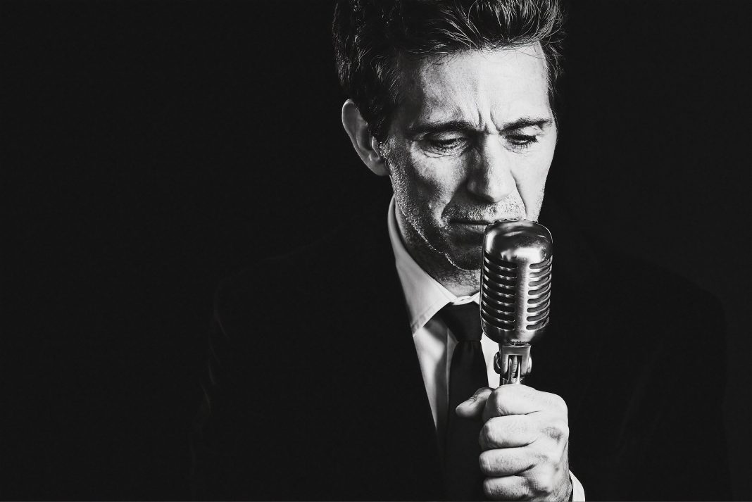 black and white photo of Ronnie Marmo in a suit with his hand on a a vintage stand microphone
