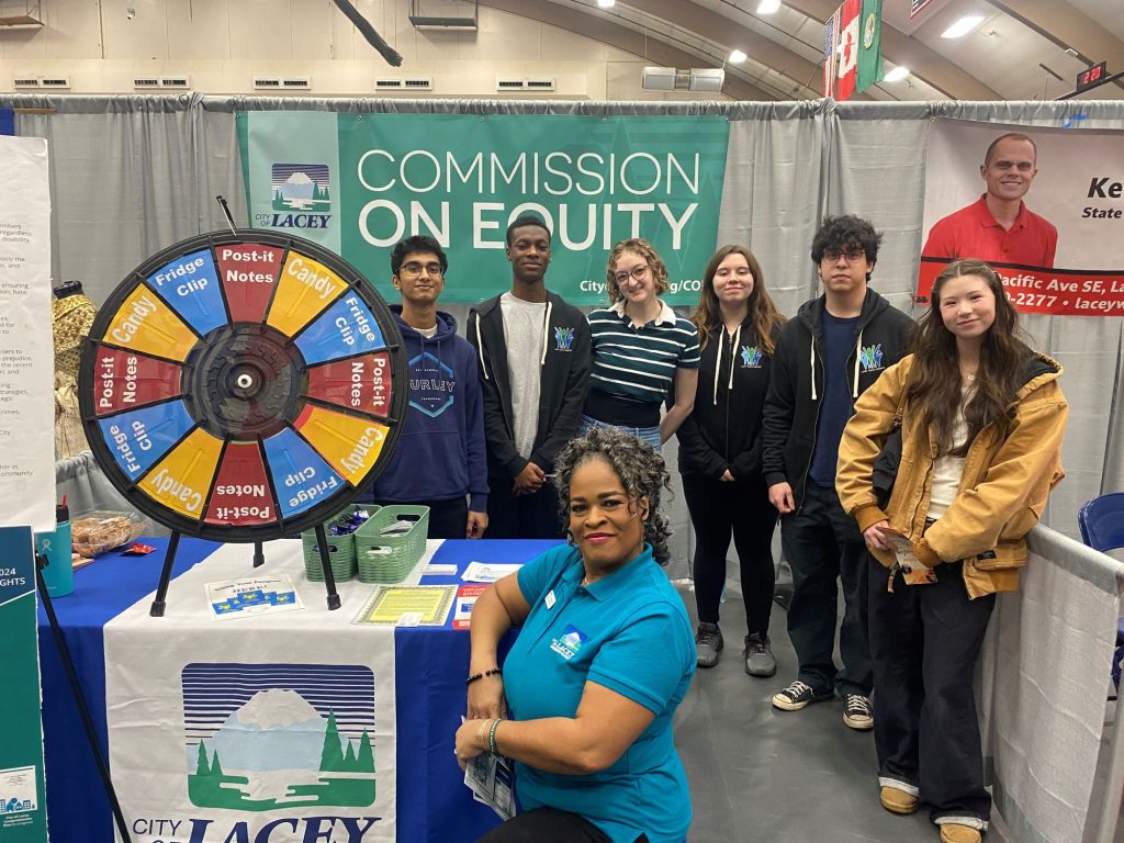6 high school students stand by a table with a prize wheel. the table has a City of Lacey banner and a woman sitting next to it