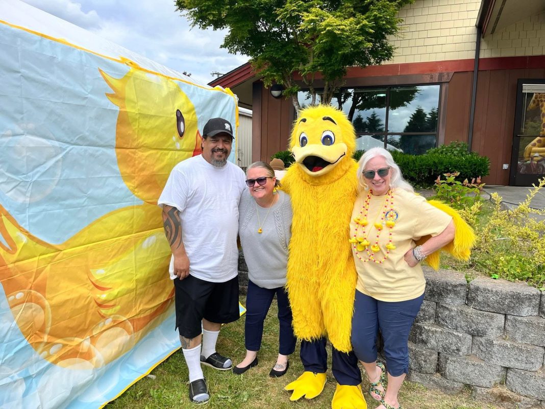 a man and two women pose for a photo with a person dressed a yellow duck costume, next to a large banner of a rubber duck at the Lacey Duck Dash