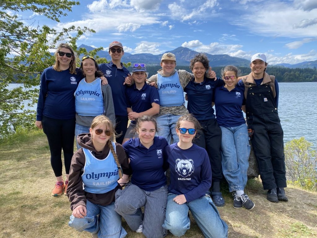 Olympia High School sailing team with Olympia Community Sailing pose for a photo outside. Three are kneeling on the ground, 8 are standing
