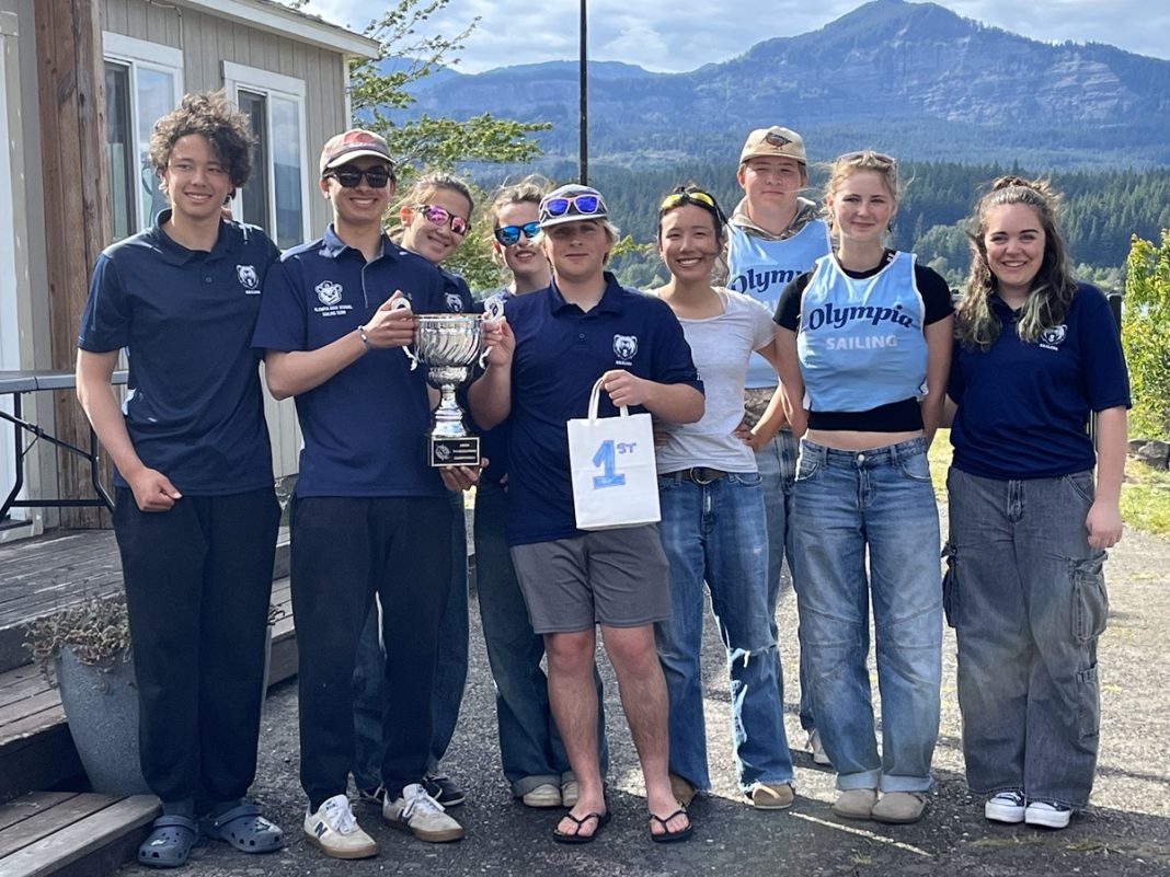 9 Olympia High School sailing team members with Olympia Community Sailing stand posed for a photo outside with their trophy and a white bag with text that says "1st" on it