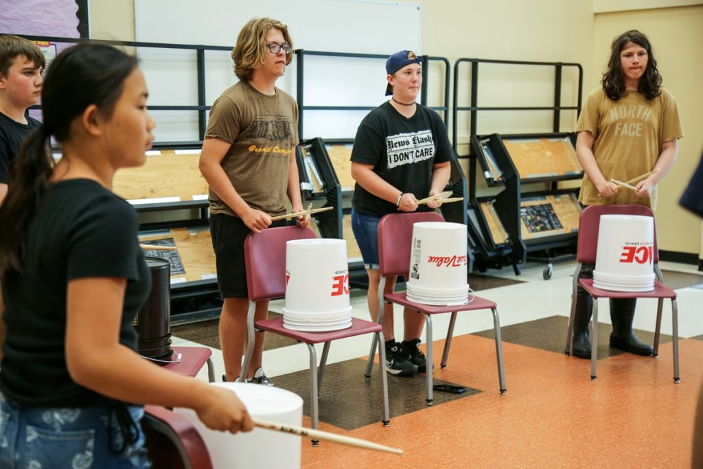 kids in a gym standing behind chairs with five-gallon buckets turned over on them, using drumsticks to beat them