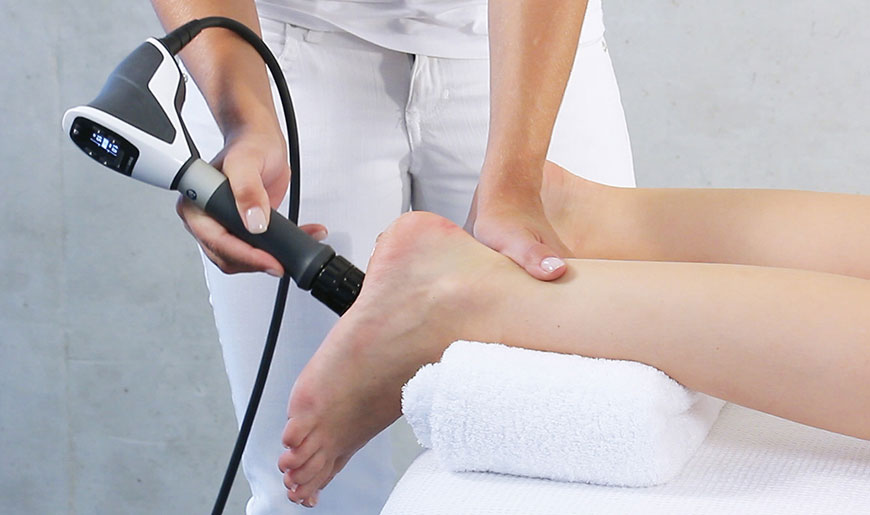 a persons feet hanging of a table resting on towels - another person stands next to them, one hand on their ankle and the other using a shockwave device on the bottom of one of the feet.